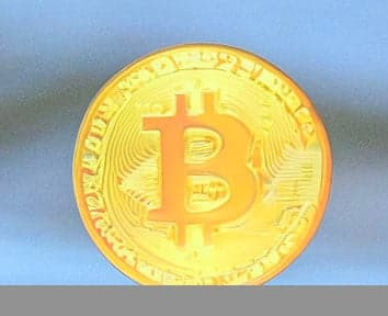 A close-up of a shiny gold coin with the Bitcoin symbol embossed on it, set against a blue background.