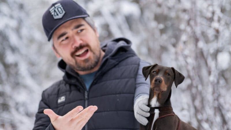 A person in a dark jacket and cap gestures animatedly while standing in a snowy landscape. A brown dog with a curious expression sits beside them.